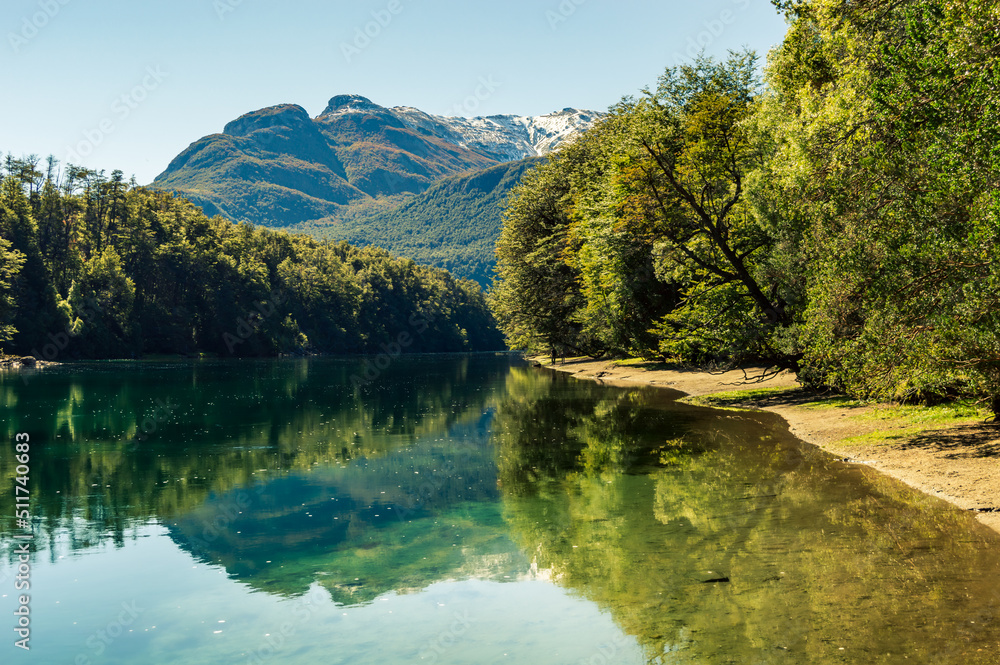 Couple walking along the coast of a river that reflects with a mountain range in the background. Arrayanes River, Los Alerces National Park.