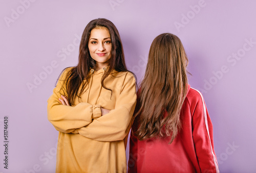 Fotografia Front view serious mother with folded arms and back view of cute daughter standing with long healthy hair in fashion hoodie together on purple studio background with empty copy space