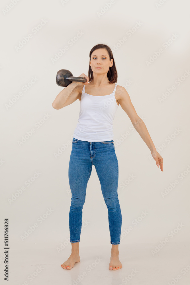 Portrait of young muscular woman wearing white top, blue jeans, standing barefoot, holding kettle bell on shoulder. Portrait of young muscular woman wearing white top, blue jeans, standing barefoot, holding kettle bell on shoulder.