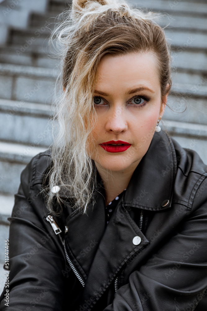 Portrait of young grey-eyed woman with long hair in bun wearing red lipstick on lips, leather jacket, sitting on step. Portrait of young grey-eyed woman with long hair in bun wearing red lipstick on lips, leather jacket, sitting on step.