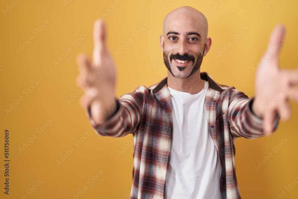 Hispanic man with beard standing over yellow background looking at the camera smiling with open arms for hug. cheerful expression embracing happiness. Hispanic man with beard standing over yellow background looking at the camera smiling with open arms for hug. cheerful expression embracing happiness.