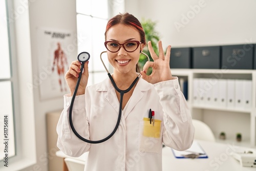 Young caucasian woman wearing doctor uniform using stethoscope doing ok sign with fingers, smiling friendly gesturing excellent symbol