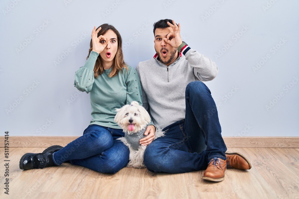 Young hispanic couple sitting on the floor with dog doing ok gesture shocked with surprised face, eye looking through fingers. unbelieving expression. Young hispanic couple sitting on the floor with dog doing ok gesture shocked with surprised face, eye looking through fingers. unbelieving expression.