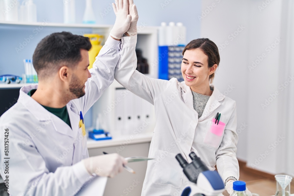 Man and woman wearing scientist uniform high five with hands raised up at laboratory Man and woman wearing scientist uniform high five with hands raised up at laboratory