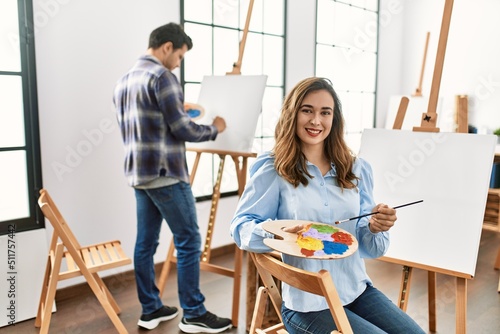 Two hispanic students painting at art school. Woman smiling happy holding paintbrushes and palette.