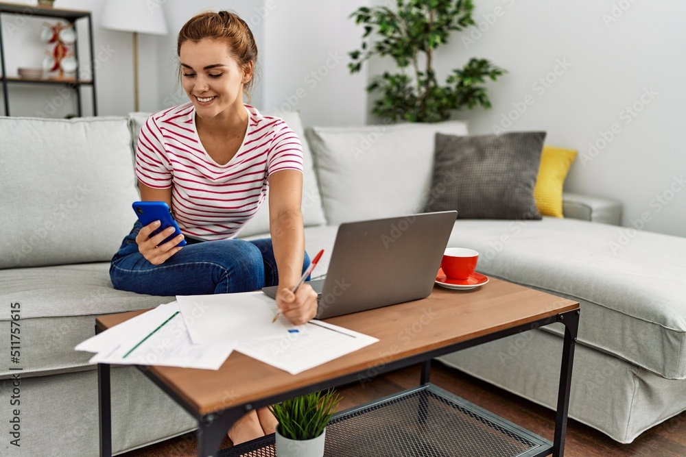 Young woman using smartphone and laptop working sitting on sofa at home Young woman using smartphone and laptop working sitting on sofa at home