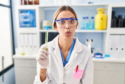 Young blonde woman working at scientist laboratory holding sample scared and amazed with open mouth for surprise  disbelief face