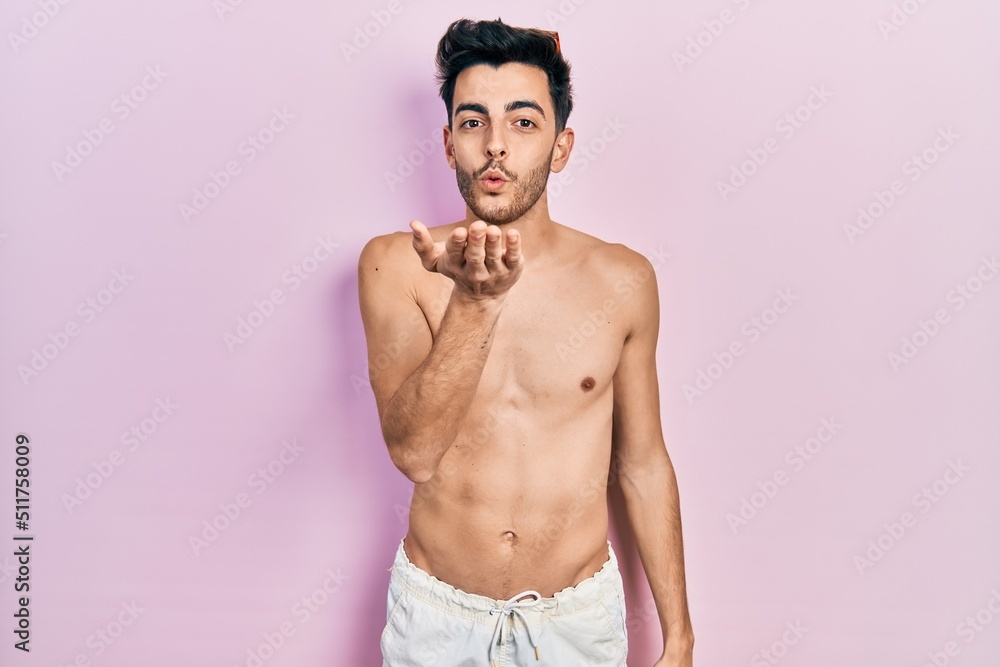 Fototapeta premium Young hispanic man wearing swimwear shirtless looking at the camera blowing a kiss with hand on air being lovely and sexy. love expression.