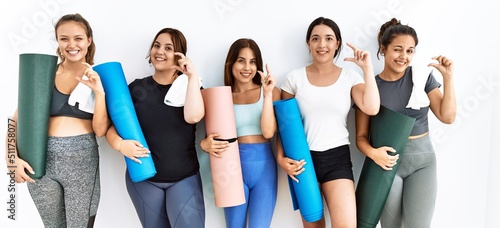 Group of women holding yoga mat standing over isolated background smiling and confident gesturing with hand doing small size sign with fingers looking and the camera. measure concept.