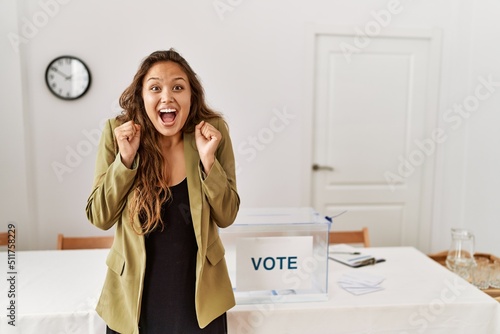 Beautiful hispanic woman standing at political campaign room celebrating crazy and amazed for success with arms raised and open eyes screaming excited. winner concept