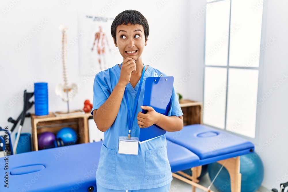 Obraz premium Young hispanic woman with short hair working at pain recovery clinic thinking worried about a question, concerned and nervous with hand on chin