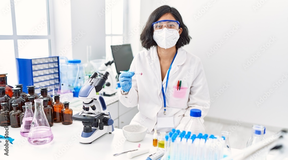 Young latin woman wearing scientist uniform and medical mask holding pill at laboratory Young latin woman wearing scientist uniform and medical mask holding pill at laboratory