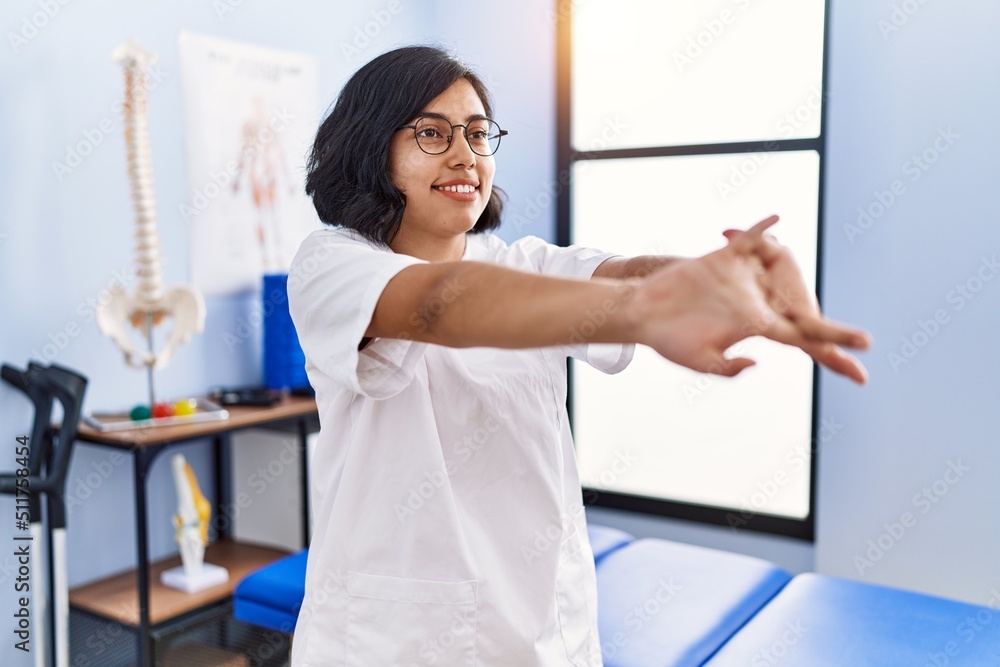 Young latin woman wearing doctor uniform stretching arms at physiotherapy clinic Young latin woman wearing doctor uniform stretching arms at physiotherapy clinic