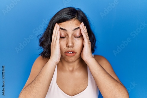 Young hispanic woman standing over blue background with hand on head for pain in head because stress. suffering migraine.
