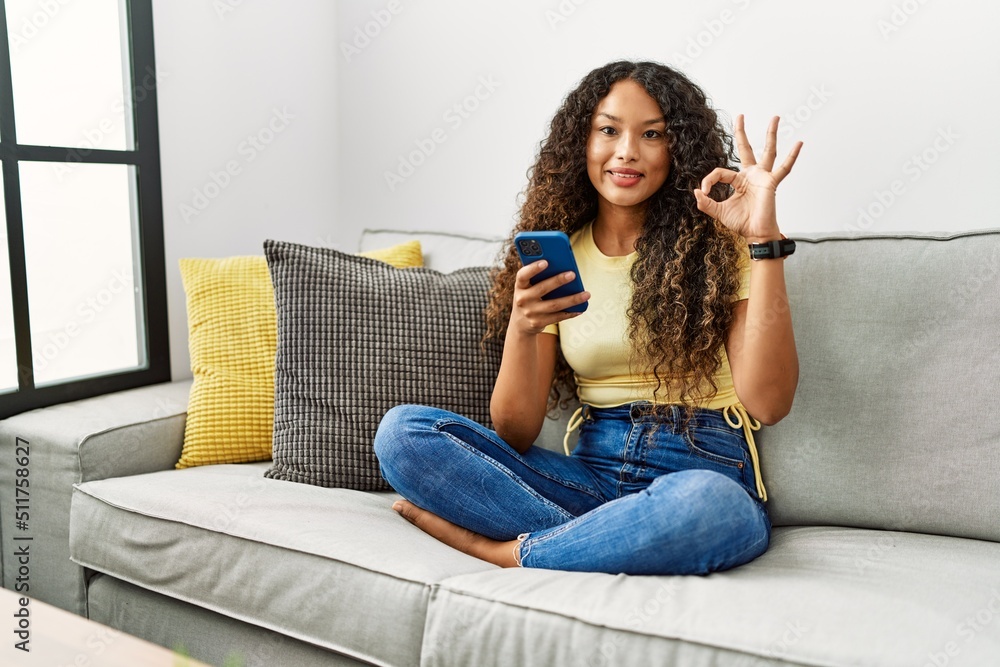 Beautiful hispanic woman sitting on the sofa at home using smartphone smiling positive doing ok sign with hand and fingers. successful expression. Beautiful hispanic woman sitting on the sofa at home using smartphone smiling positive doing ok sign with hand and fingers. successful expression.