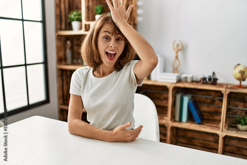 Young caucasian girl wearing casual clothes sitting on the table at home surprised with hand on head for mistake, remember error. forgot, bad memory concept. Young caucasian girl wearing casual clothes sitting on the table at home surprised with hand on head for mistake, remember error. forgot, bad memory concept.