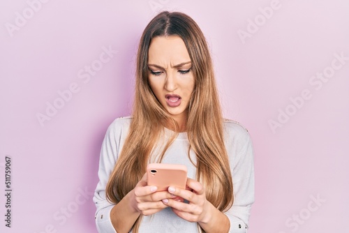 Young blonde girl using smartphone typing message in shock face, looking skeptical and sarcastic, surprised with open mouth