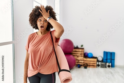African american woman with afro hair holding yoga mat at pilates room surprised with hand on head for mistake, remember error. forgot, bad memory concept. photo