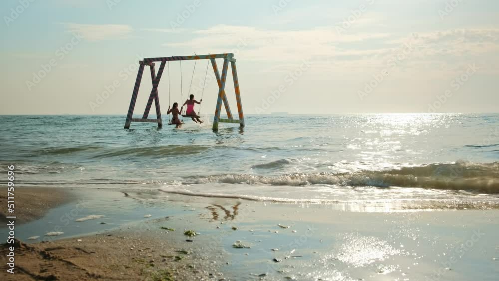 Young women on the swing in the sea. Two happy girls on the beach swinging in the blue water, summer vacation. Young women on the swing in the sea. Two happy girls on the beach swinging in the blue water, summer vacation.