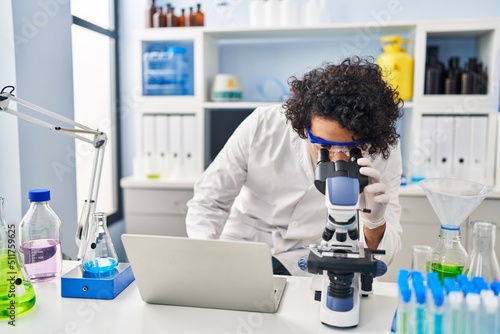 Young hispanic man wearing scientist uniform using laptop and microscope at laboratory