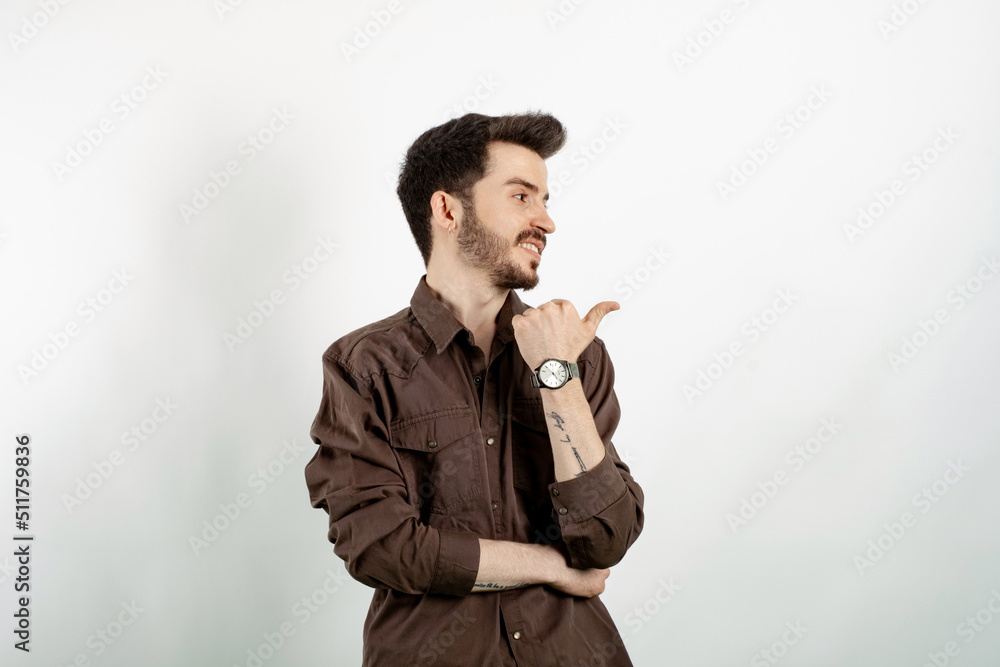 Handsome young man wearing casual clothes posing isolated over white background looking and pointing to the side with thumb up. Handsome young man wearing casual clothes posing isolated over white background looking and pointing to the side with thumb up.
