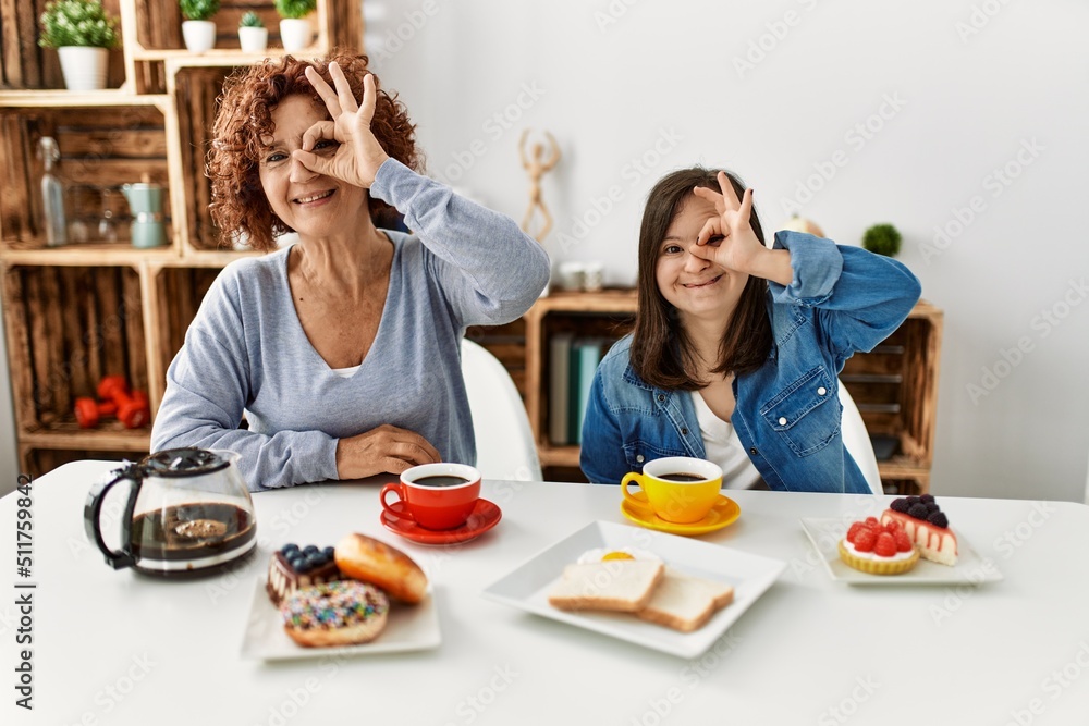 Family of mother and down syndrome daughter sitting at home eating breakfast doing ok gesture with hand smiling, eye looking through fingers with happy face. Family of mother and down syndrome daughter sitting at home eating breakfast doing ok gesture with hand smiling, eye looking through fingers with happy face.