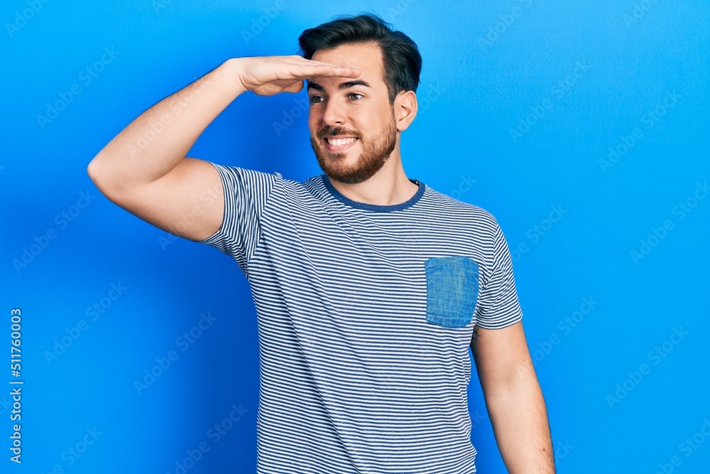 Handsome caucasian man with beard wearing casual striped t shirt very happy and smiling looking far away with hand over head. searching concept. Handsome caucasian man with beard wearing casual striped t shirt very happy and smiling looking far away with hand over head. searching concept.