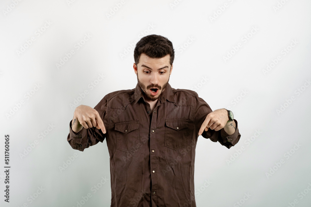Naklejka premium Handsome young man wearing casual clothes posing isolated over white background surprised and shouting with pointing bottom with his hand. Give advice, recommend click link, check-out product.