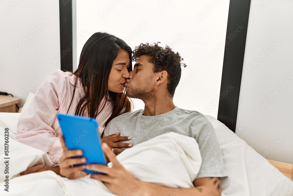 Young latin couple kissing and using touchpad lying on the bed at bedroom. Young latin couple kissing and using touchpad lying on the bed at bedroom.