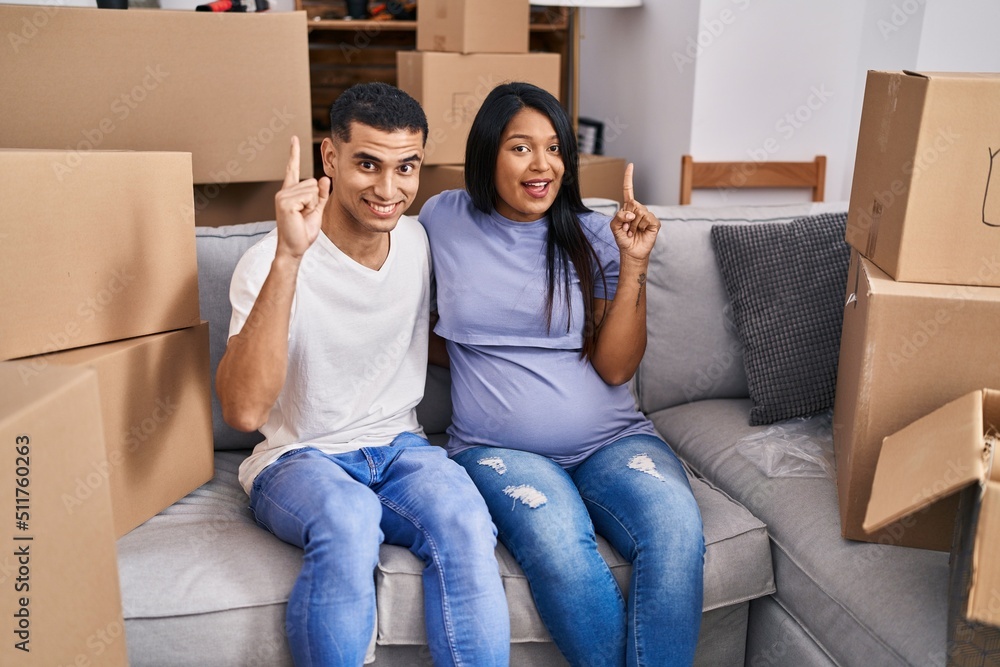 Young hispanic couple expecting a baby sitting on the sofa at new home smiling with an idea or question pointing finger with happy face, number one Young hispanic couple expecting a baby sitting on the sofa at new home smiling with an idea or question pointing finger with happy face, number one