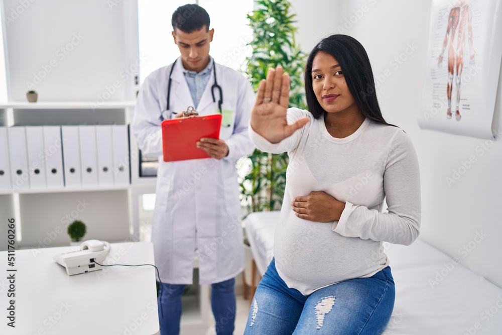 Fototapeta premium Young hispanic mother expecting a baby at the doctor with open hand doing stop sign with serious and confident expression, defense gesture