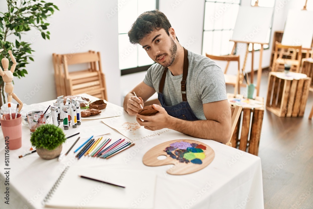 Young hispanic artist man smiling happy painting pottery at art studio.