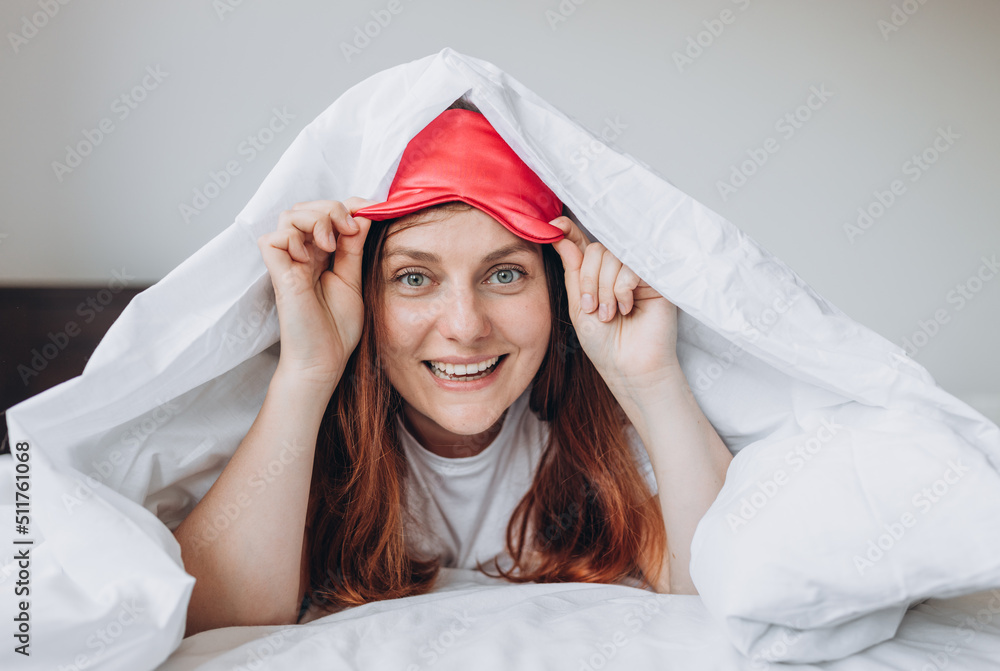 Cheerful young happy woman in pajama smiling and lifting sleep mask after awakening in morning. Redhead girl waking up enjoy good morning looking at camera Cheerful young happy woman in pajama smiling and lifting sleep mask after awakening in morning. Redhead girl waking up enjoy good morning looking at camera