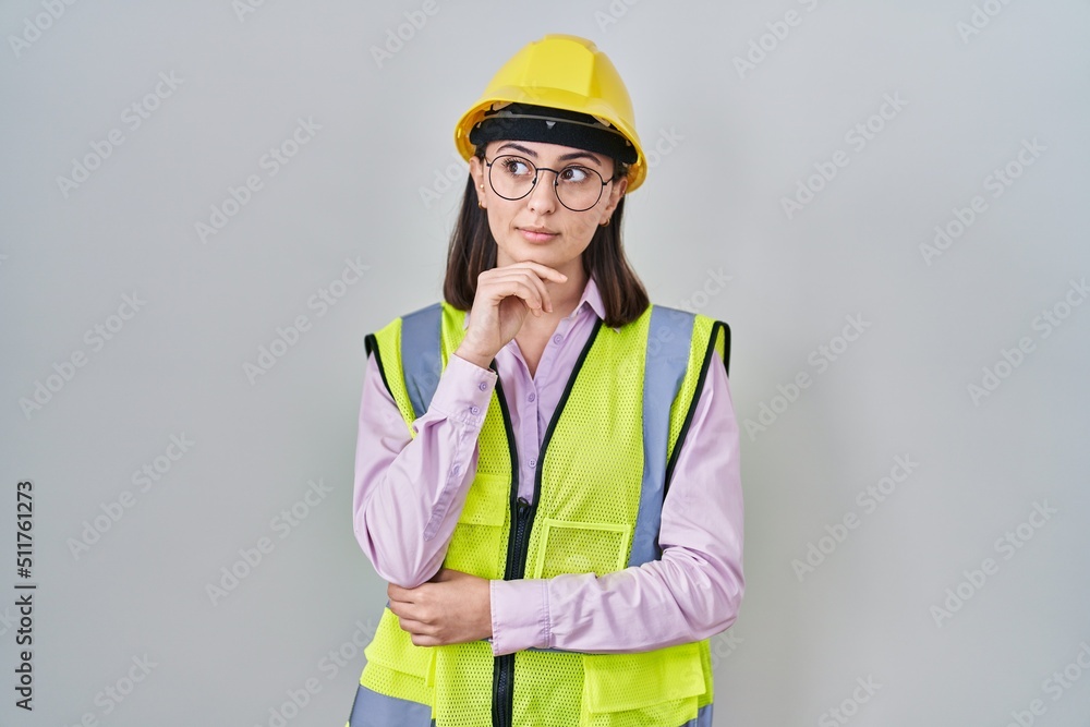 Hispanic girl wearing builder uniform and hardhat with hand on chin thinking about question, pensive expression. smiling with thoughtful face. doubt concept.