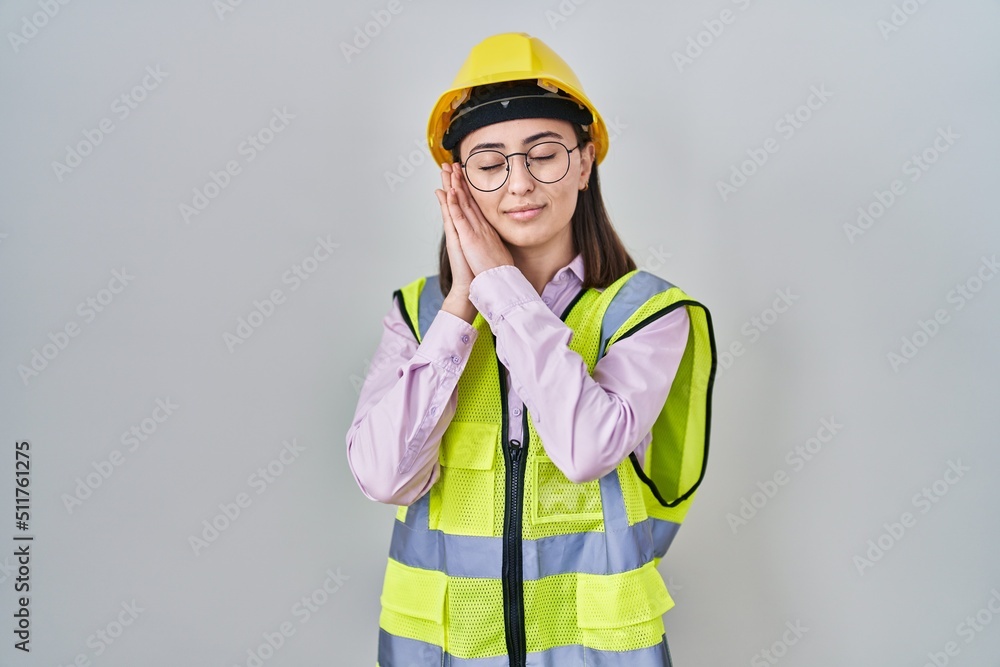 Hispanic girl wearing builder uniform and hardhat sleeping tired dreaming and posing with hands together while smiling with closed eyes. Hispanic girl wearing builder uniform and hardhat sleeping tired dreaming and posing with hands together while smiling with closed eyes.