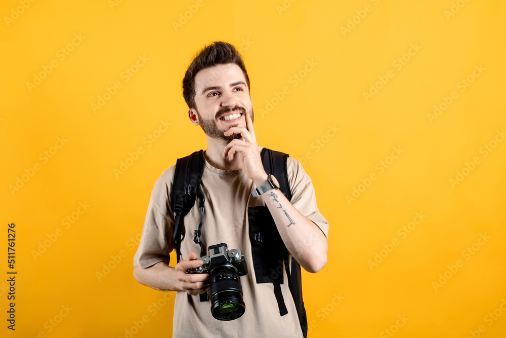 Young man smiling confident wearing beige tee posing isolated over yellow background curious dreamy looking empty space smiling touching chin while holding a dslr camera. Young man smiling confident wearing beige tee posing isolated over yellow background curious dreamy looking empty space smiling touching chin while holding a dslr camera.