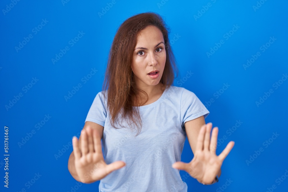 Fototapeta premium Brunette woman standing over blue background moving away hands palms showing refusal and denial with afraid and disgusting expression. stop and forbidden.