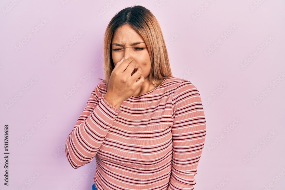 Beautiful hispanic woman wearing casual striped sweater smelling something stinky and disgusting, intolerable smell, holding breath with fingers on nose. bad smell Beautiful hispanic woman wearing casual striped sweater smelling something stinky and disgusting, intolerable smell, holding breath with fingers on nose. bad smell