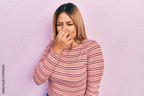 Beautiful hispanic woman wearing casual striped sweater smelling something stinky and disgusting, intolerable smell, holding breath with fingers on nose. bad smell
