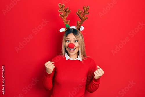 Beautiful hispanic woman wearing deer christmas hat and red nose celebrating surprised and amazed for success with arms raised and open eyes. winner concept.