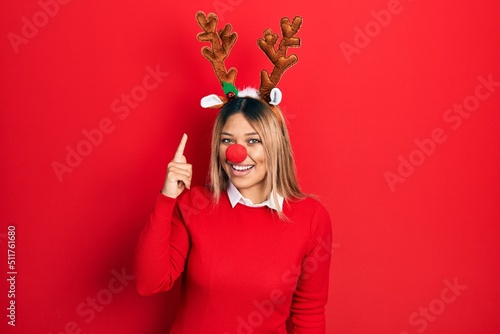 Beautiful hispanic woman wearing deer christmas hat and red nose pointing finger up with successful idea. exited and happy. number one.
