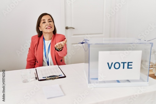 Beautiful middle age hispanic woman at political election sitting by ballot smiling cheerful offering palm hand giving assistance and acceptance.