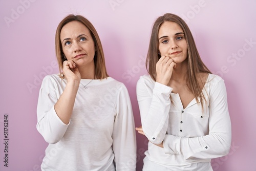 Middle age mother and young daughter standing over pink background with hand on chin thinking about question, pensive expression. smiling with thoughtful face. doubt concept.