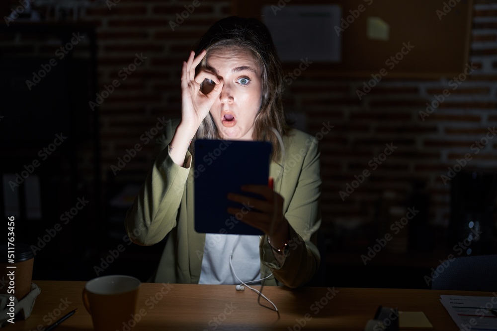 Blonde caucasian woman working at the office at night doing ok gesture shocked with surprised face, eye looking through fingers. unbelieving expression. Blonde caucasian woman working at the office at night doing ok gesture shocked with surprised face, eye looking through fingers. unbelieving expression.