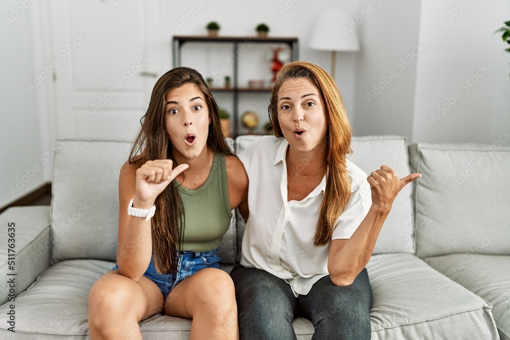 Mother and daughter together sitting on the sofa at home surprised pointing with hand finger to the side, open mouth amazed expression.
