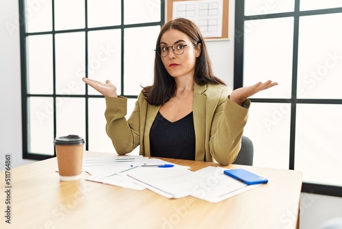 Young brunette woman wearing business style sitting on desk at office clueless and confused expression with arms and hands raised. doubt concept.