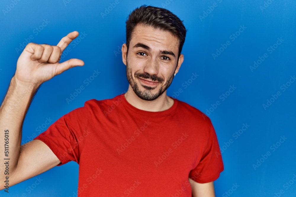 Young hispanic man with beard wearing red t shirt over blue background smiling and confident gesturing with hand doing small size sign with fingers looking and the camera. measure concept. Young hispanic man with beard wearing red t shirt over blue background smiling and confident gesturing with hand doing small size sign with fingers looking and the camera. measure concept.