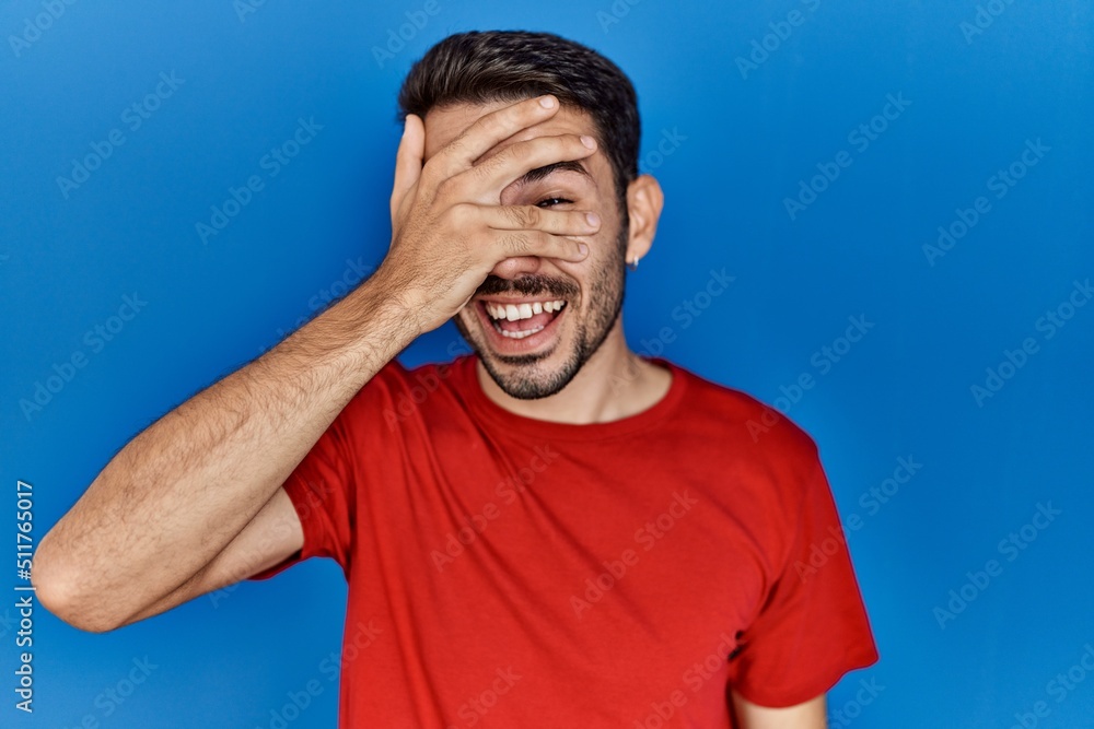 Young hispanic man with beard wearing red t shirt over blue background peeking in shock covering face and eyes with hand, looking through fingers with embarrassed expression. Young hispanic man with beard wearing red t shirt over blue background peeking in shock covering face and eyes with hand, looking through fingers with embarrassed expression.