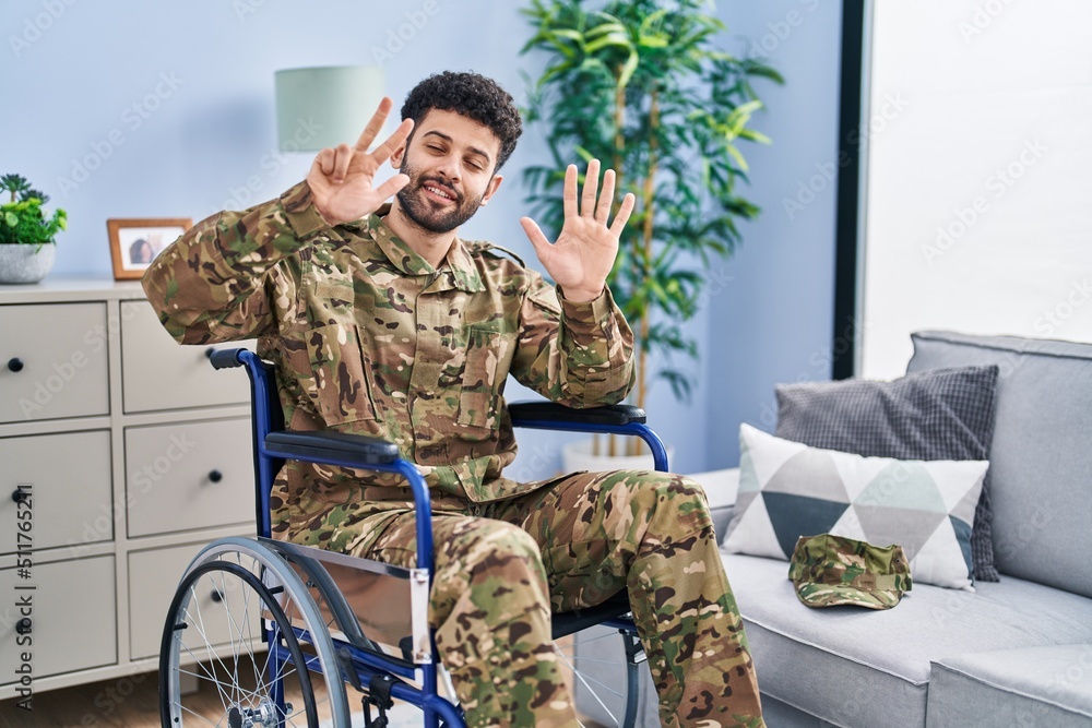 Arab man wearing camouflage army uniform sitting on wheelchair showing and pointing up with fingers number eight while smiling confident and happy.