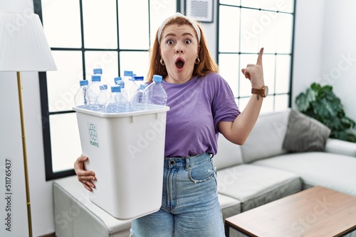 Young redhead woman holding recycling wastebasket with plastic bottles pointing finger up with successful idea. exited and happy. number one.